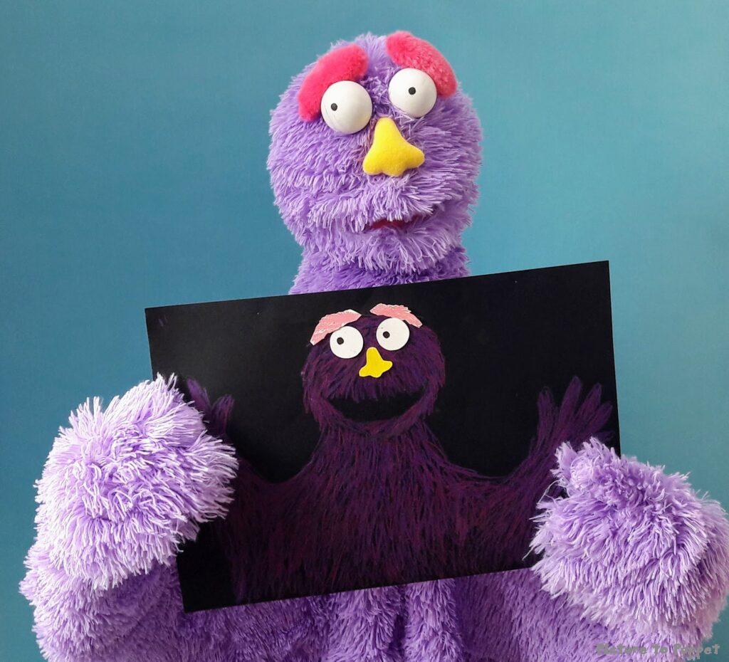 A purple puppet holding up a puppet design showing the progression from picture to puppet.