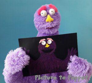 A purple puppet holding up a puppet design showing the progression from picture to puppet.